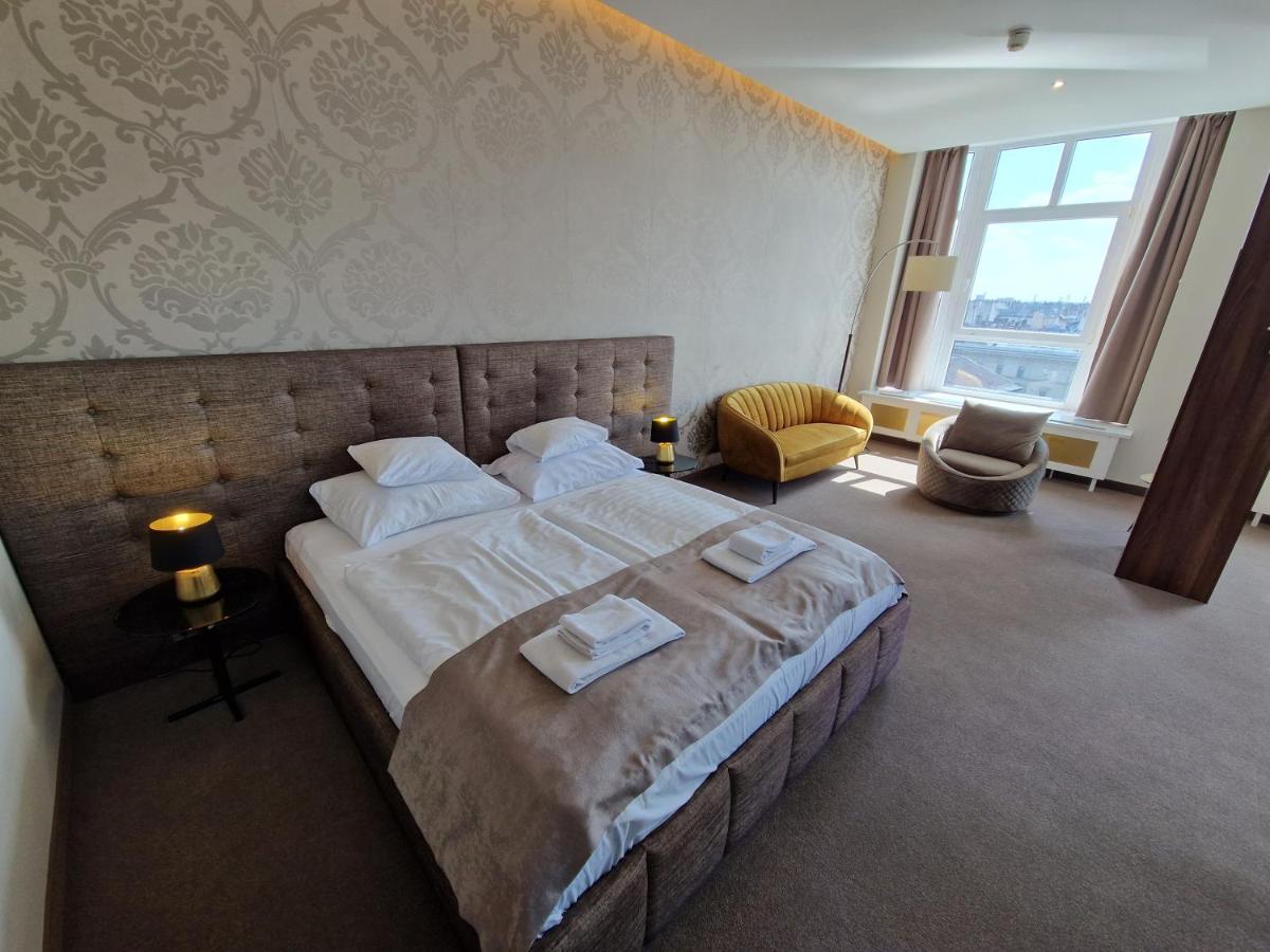 hotel room with beige bed, patterned damask wallpaper in beige, yellow sofa and large window with views of Budapest